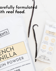 White packaging of French Vanilla protein powder with a stick pack and vanilla beans on a light surface.