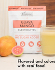 Pouch of Guava Mango Electrolytes next to a whole mango, a halved guava, and a glass of yellow liquid.