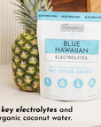 Pouch of Blue Hawaiian Electrolytes next to a pineapple, featuring details about ingredients and servings.