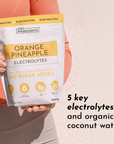 A person holds a pouch of Orange Pineapple Electrolytes with a white and yellow design, featuring text about its ingredients.