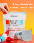 Hand holding Rocket Berry Electrolytes packet with red and blue gradient design, displayed with popsicle and glass of water o