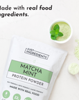 White pouch of matcha mint protein powder with green label, surrounded by green powder and a bowl of mixed powder topped with