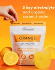 White pouch labeled "Orange Electrolytes" with orange banner, surrounded by fresh orange slices and a glass of orange-colored