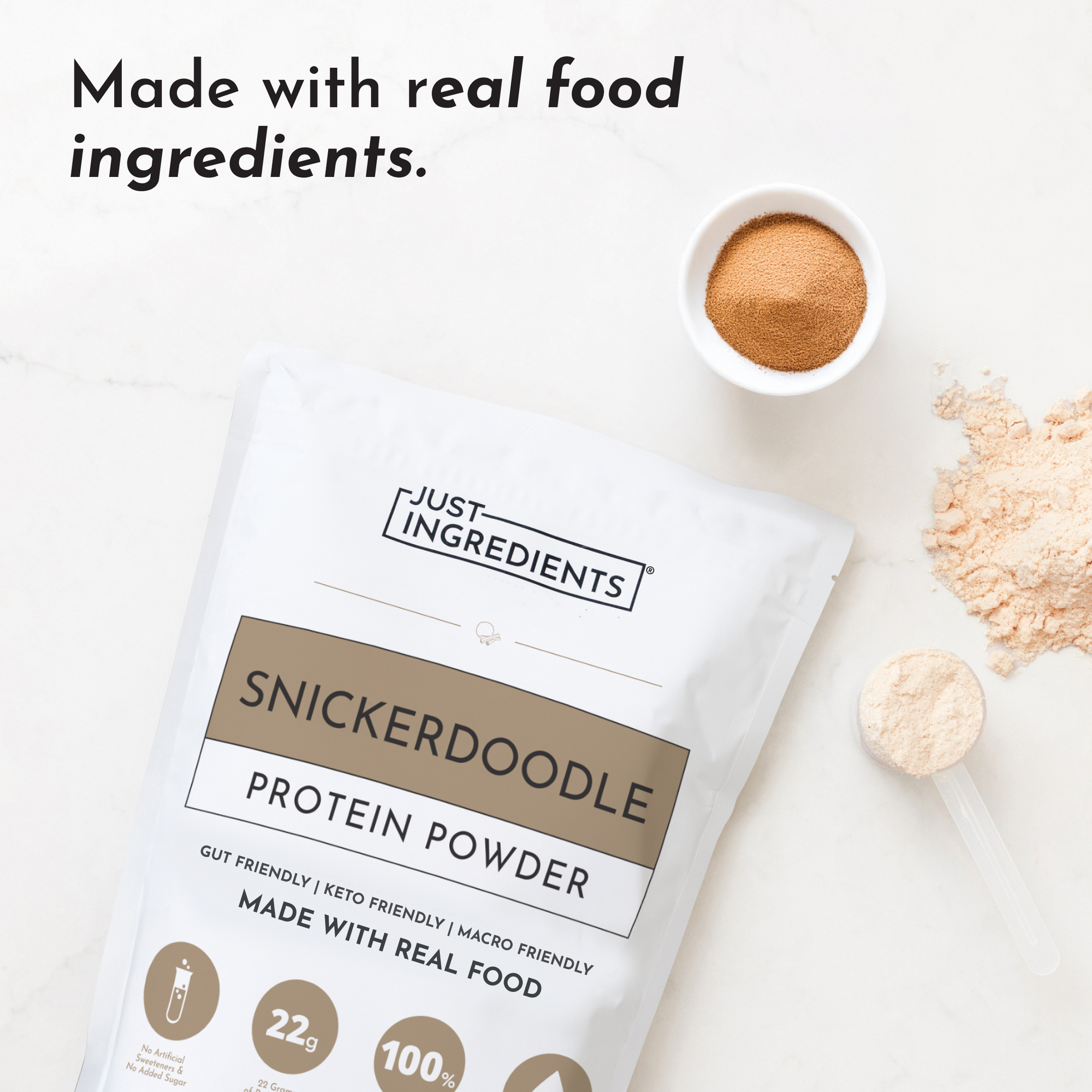 Snickerdoodle protein powder pouch with cinnamon powder in white bowl and measuring scoops on white surface.
