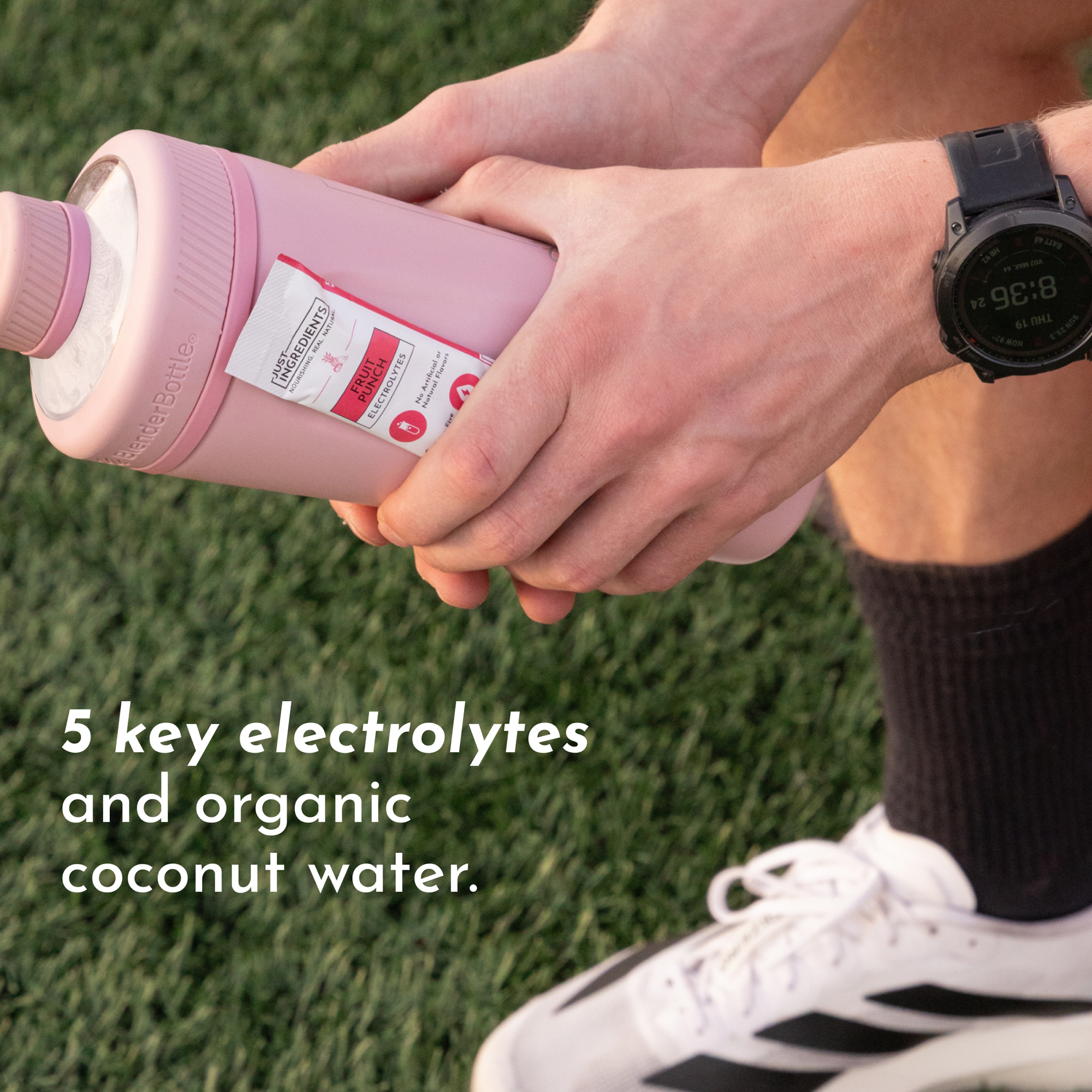A person holds a pink sports bottle with a label indicating fruit punch flavor and electrolytes, on grass.