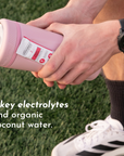A person holds a pink sports bottle with a label indicating fruit punch flavor and electrolytes, on grass.