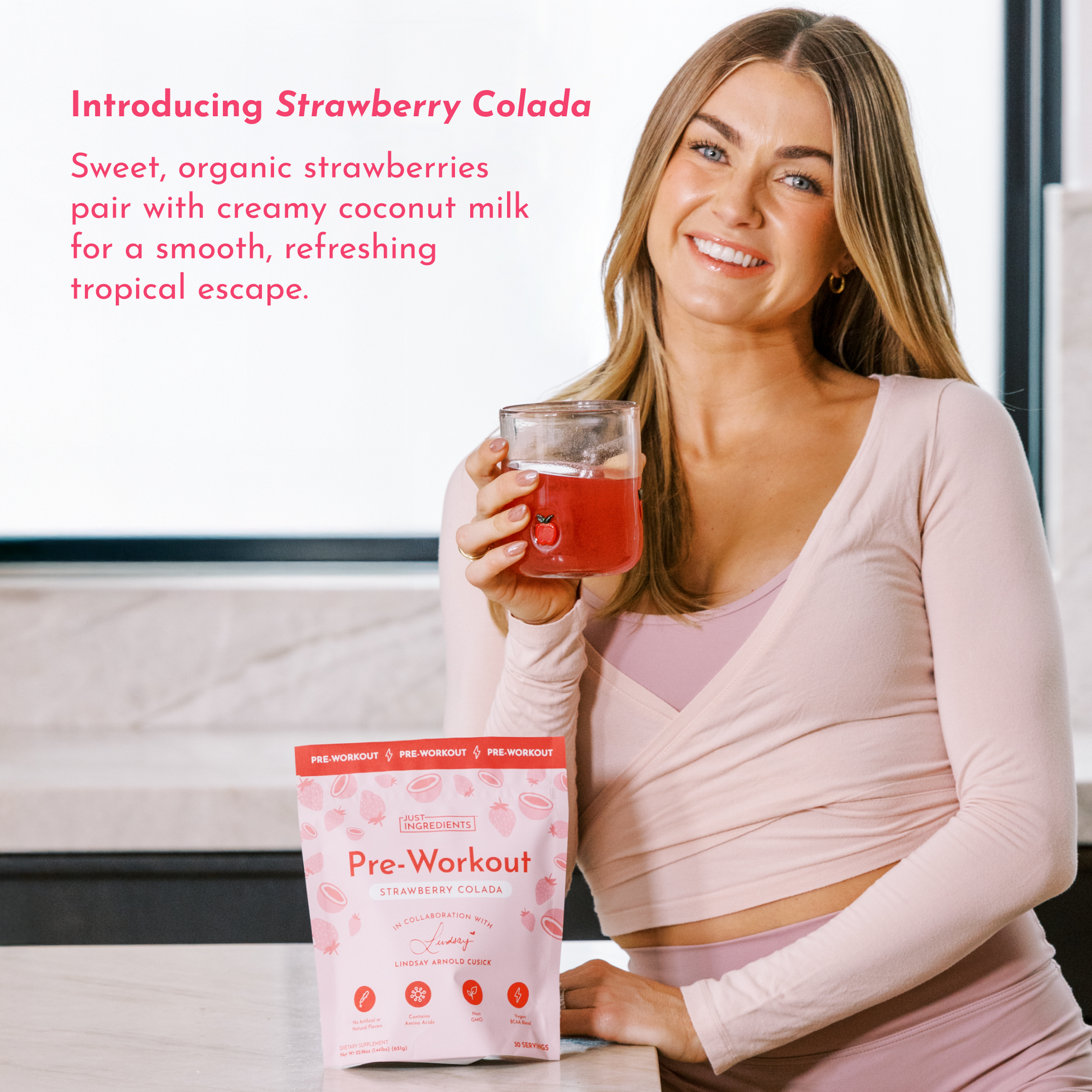 Woman holding a glass of pink drink next to a bag of Strawberry Colada Pre-Workout on a countertop.