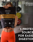 A person holds a bag of creamy peanut butter chocolate protein powder in a gym setting.
