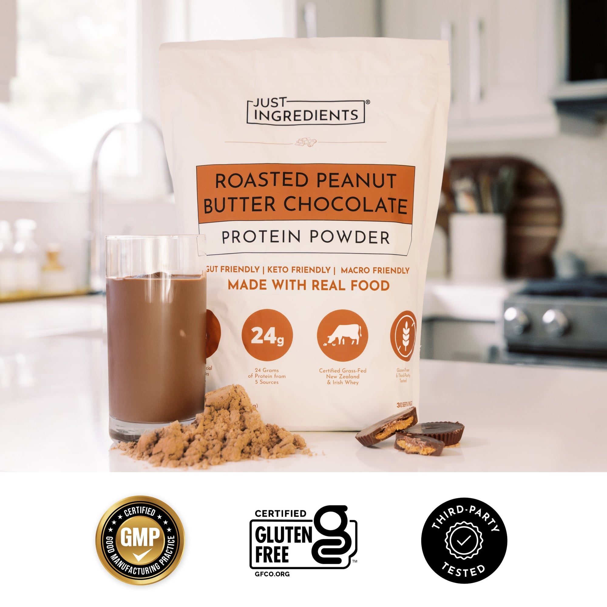 Hand holding a glass of chocolate protein shake next to a bag of Roasted Peanut Butter Chocolate Protein Powder and shelled peanuts.