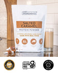 Salted caramel protein bag next to a prepared protein shake and a pink handheld mixer on a countertop.