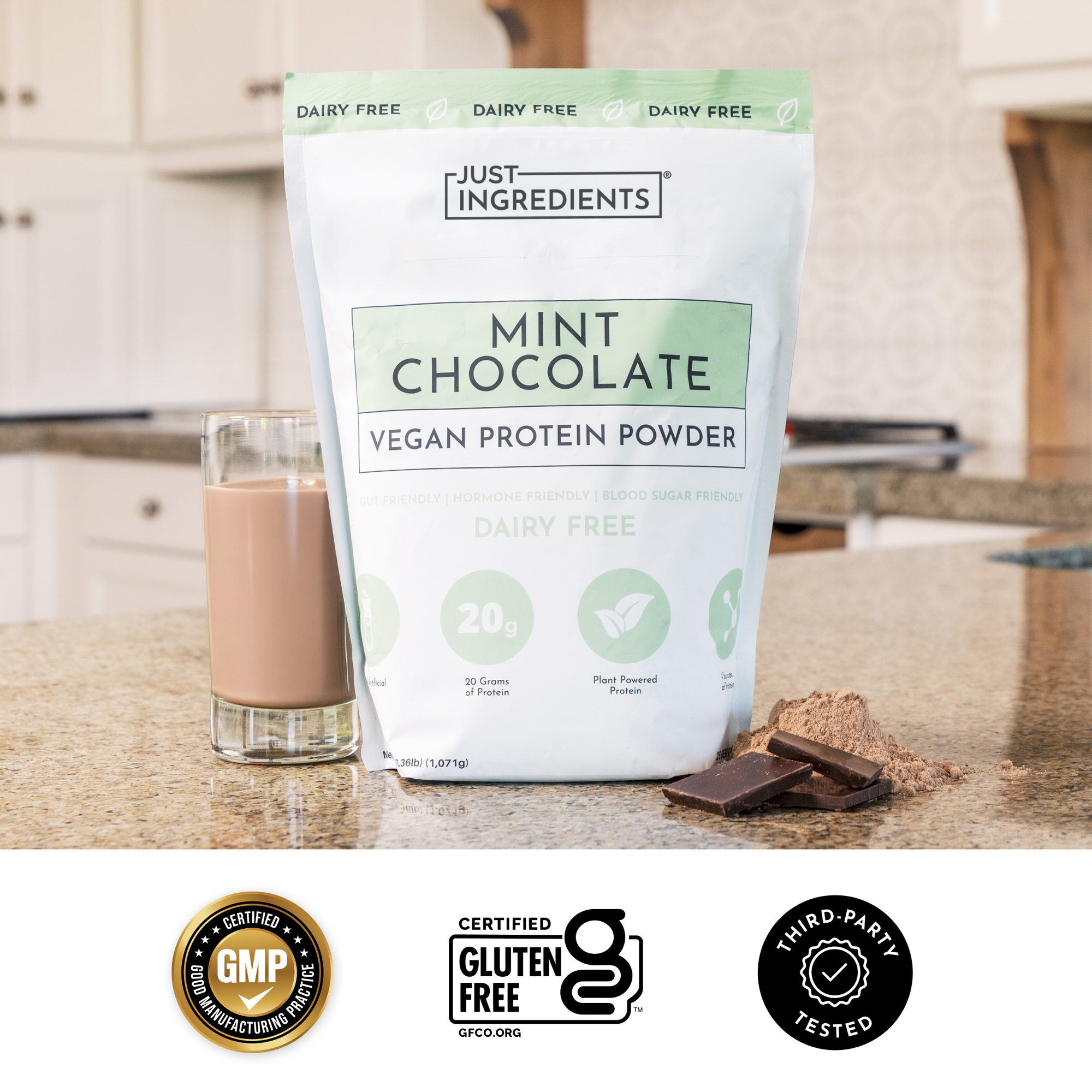 Clear scoop of beige protein powder held above a glass with pieces of dark chocolate in the background.
