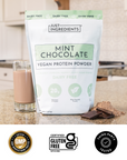 Clear scoop of beige protein powder held above a glass with pieces of dark chocolate in the background.
